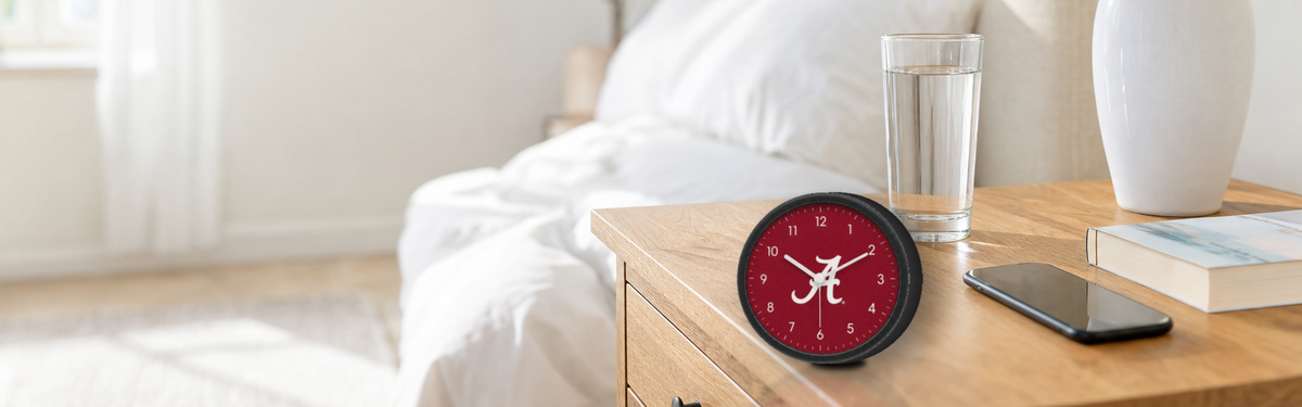 A small crimson team clock on a bedside nightstand next to a glass of water and a phone.
